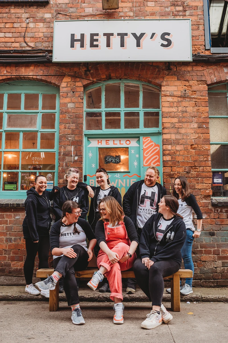 A group of eight people posing together on a wooden bench in front of a brick building with a turquoise door and a white sign reading 'HETTY'S'. 