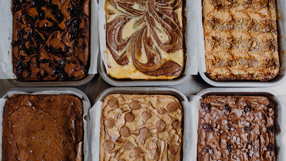 Six different types of baked brownies arranged in square metal pans lined with parchment paper, shown from a top-down view.