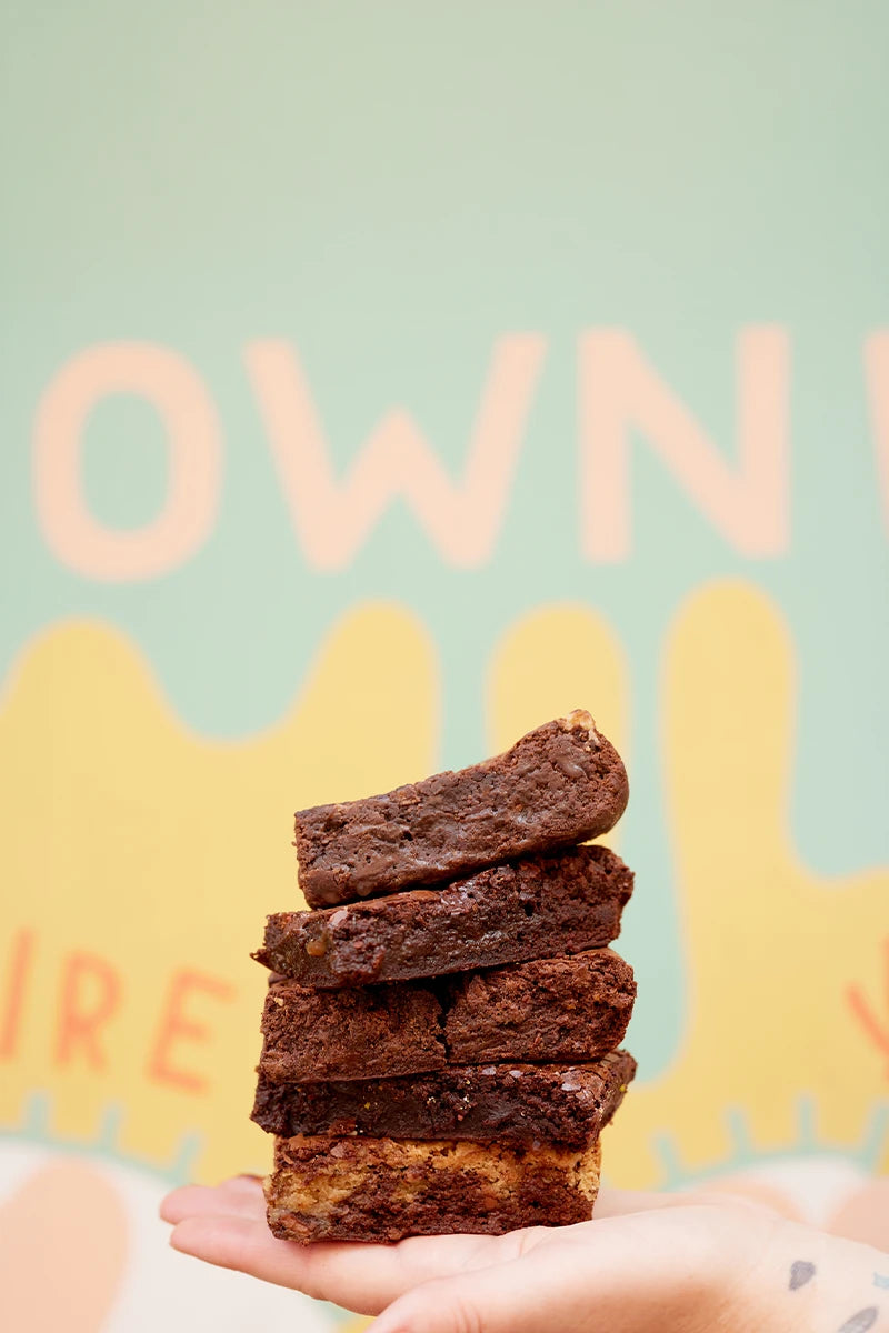 A hand holding a stack of five rich, glossy chocolate brownies in front of a colorful, blurred background with pastel text and shapes.