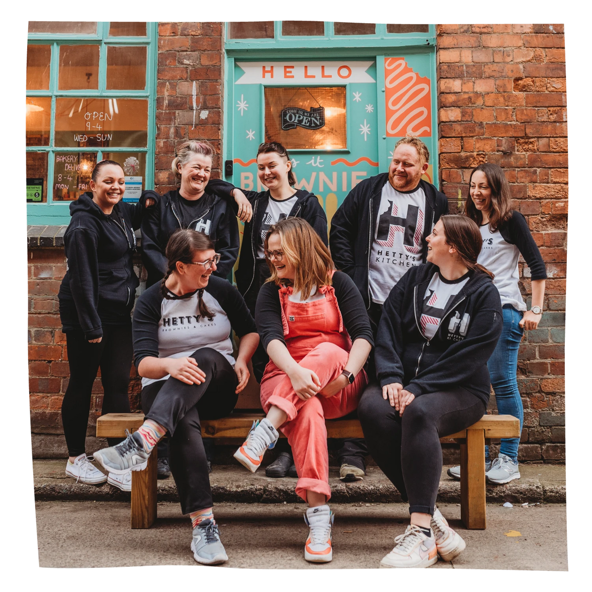 A group of eight people posing together on a wooden bench in front of a brick building with a teal door.
