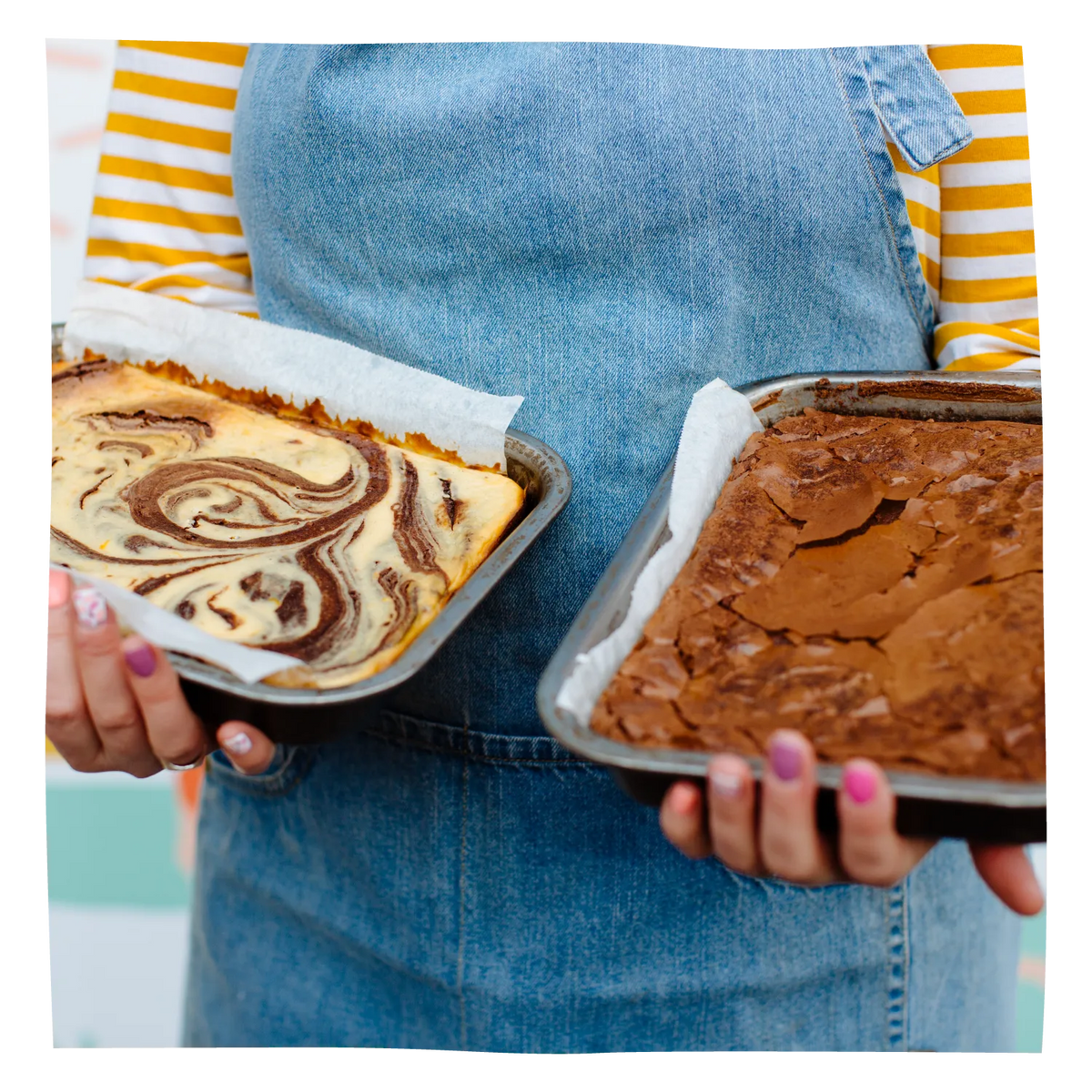 A person wearing denim overalls over a yellow-and-white striped shirt holds two baking pans of brownies. 
