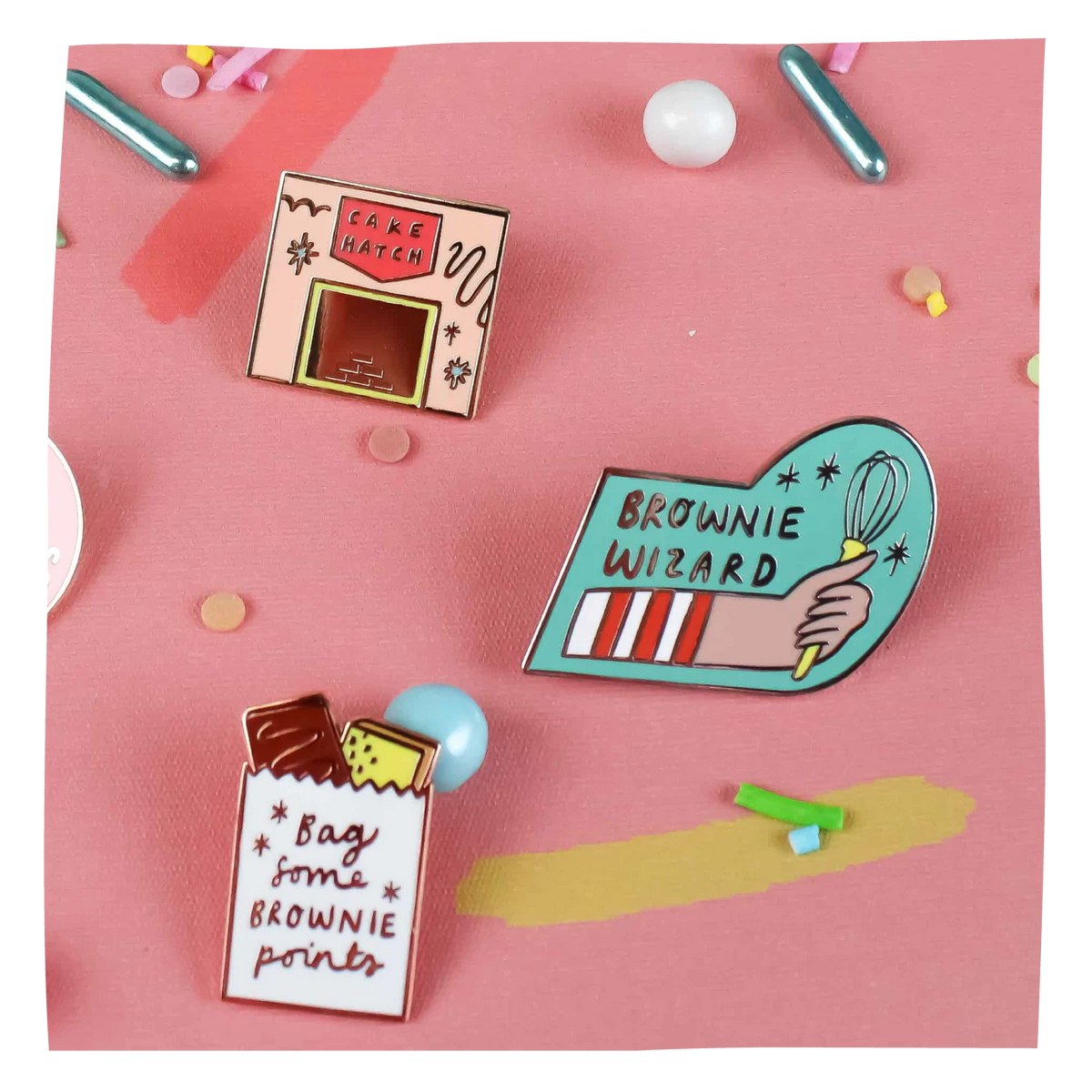 Three enamel pin badges with brownie-themed designs arranged on a pink background.