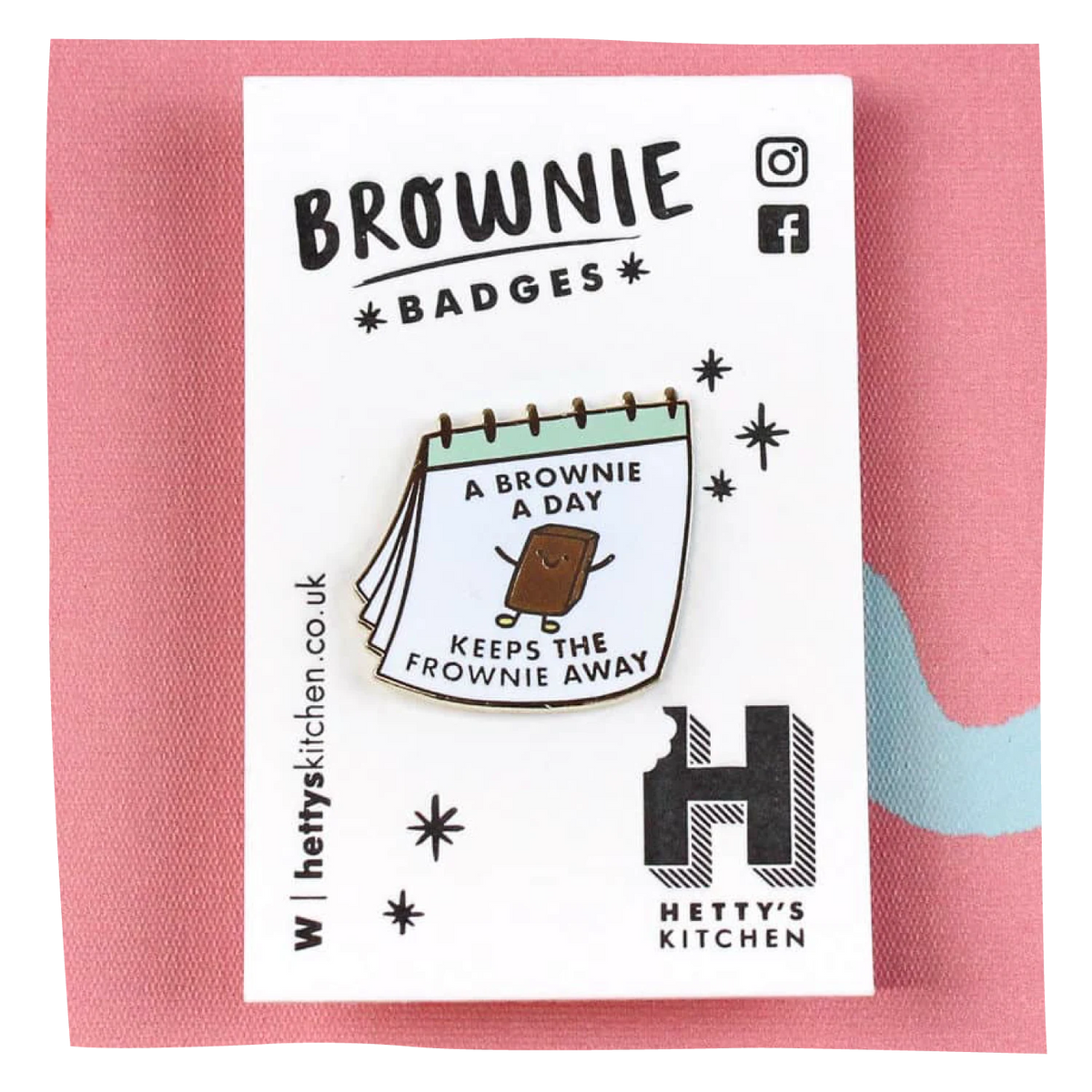 A brownie-themed enamel pin displayed on its white branded backing card, set against a pink textured fabric background. The pin features a brownie character with the text 'A BROWNIE A DAY KEEPS THE FROWNIE AWAY'.