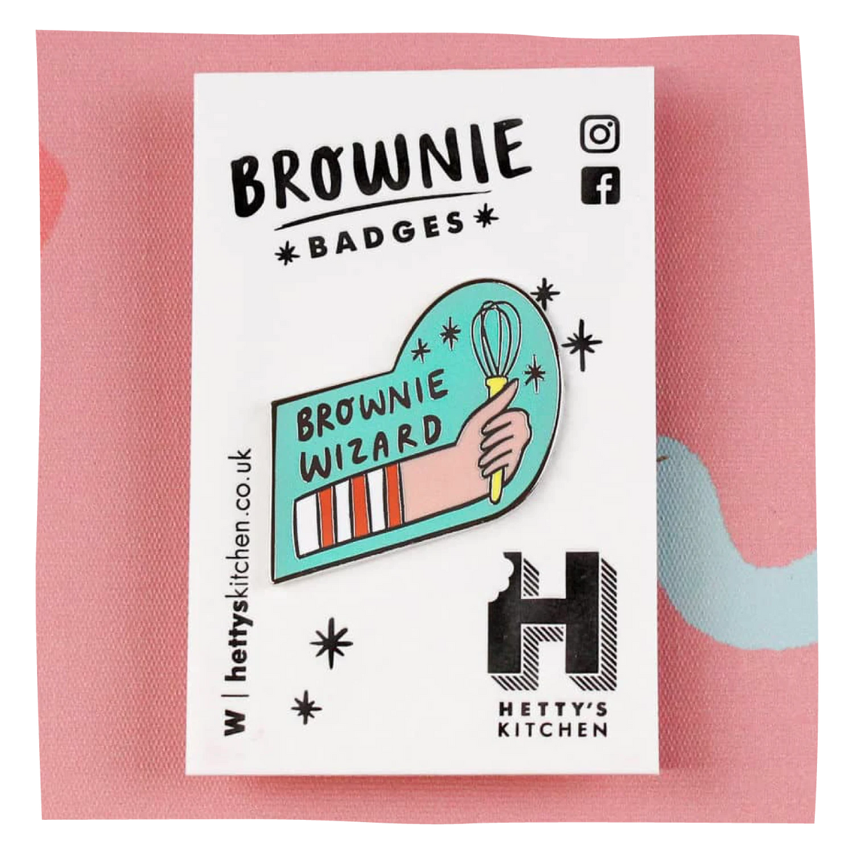 A n enamel pin featuring a hand holding a whisk, with the text 'BROWNIE WIZARD' and a striped brownie bar, displayed on a white backing card labeled 'BROWNIE BADGES' and 'HETTY'S KITCHEN'. The card is placed on a pink textured background.