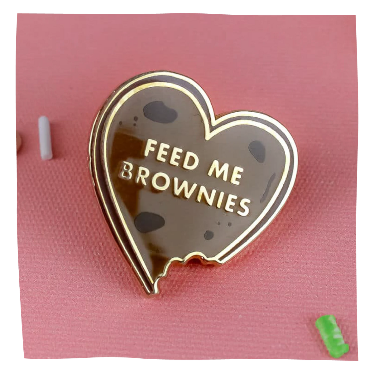 A heart-shaped enamel pin designed to look like a brownie cookie with dark chocolate chips and the words 'FEED ME BROWNIES' in gold lettering, displayed on a pink textured background.