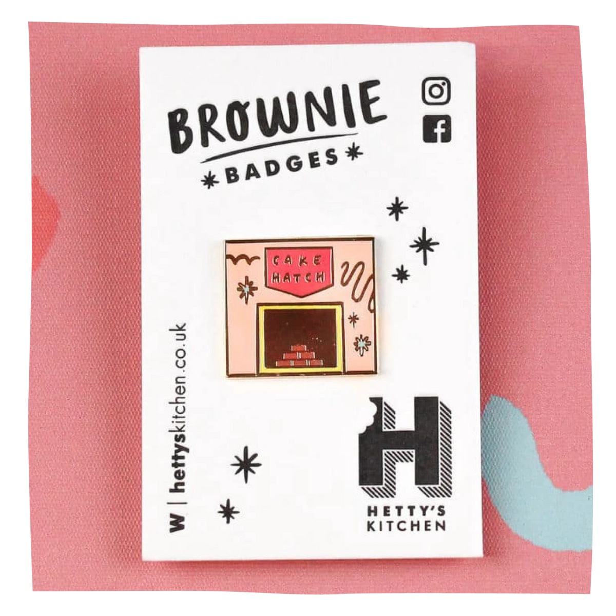 A small enamel pin shaped like a bakery or cake hatch, displayed on a white backing card labeled 'BROWNIE BADGES' with the brand 'Hetty's Kitchen' visible. The pin has pink, cream, and red details, set against a pink textured background.