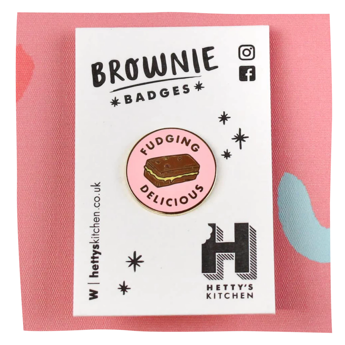 A round enamel pin featuring a brownie with the words 'FUDGING DELICIOUS' on a pink background, mounted on a white branded backing card that reads 'BROWNIE BADGES' and shows the Hetty's Kitchen logo.