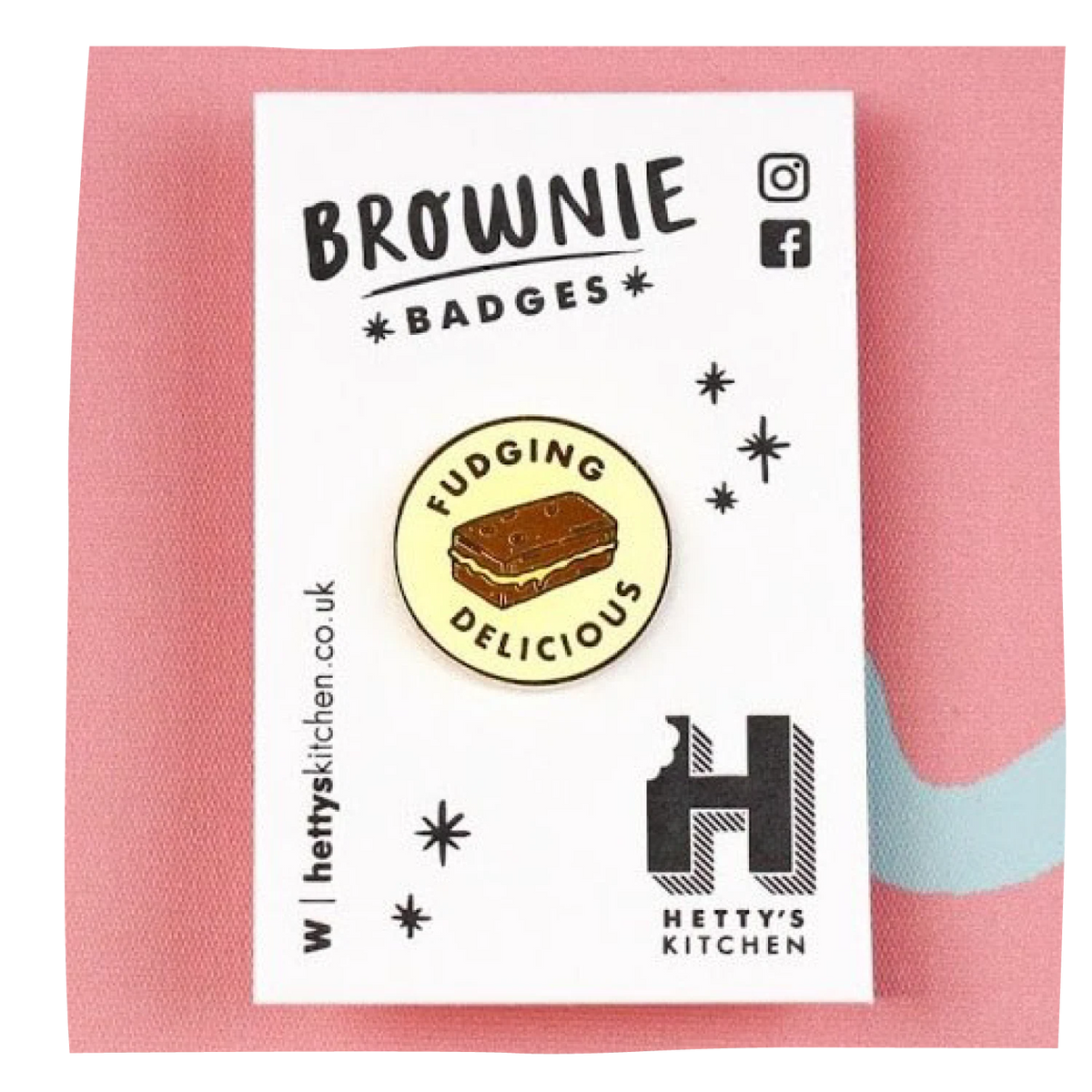 A brownie badge pin displayed on its white backing card, which features the brand name 'Hetty's Kitchen' and social media icons, set against a pink textured background.