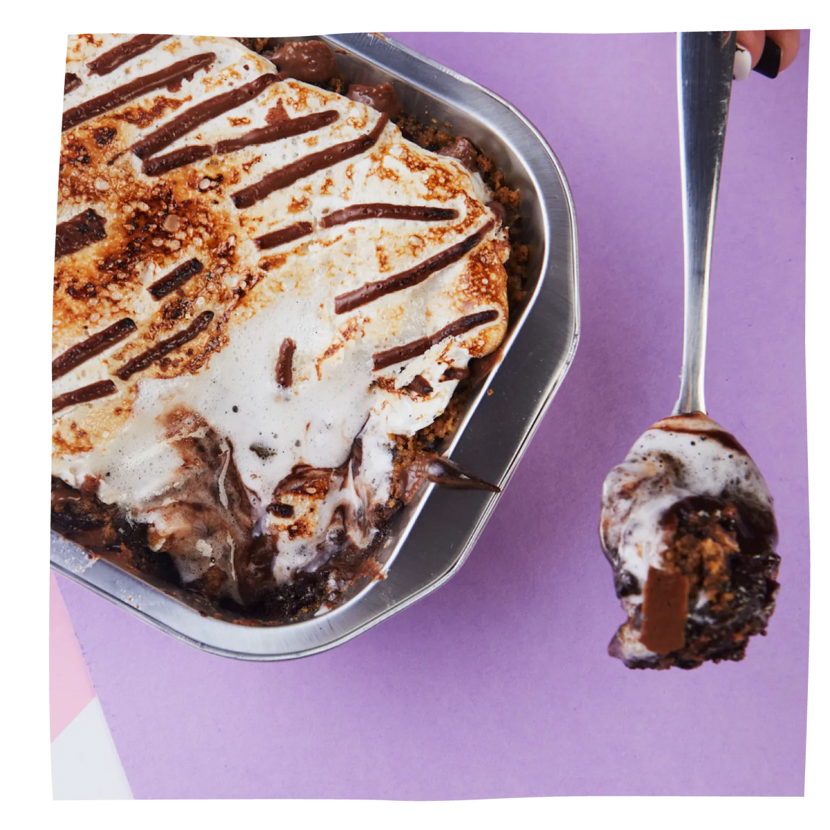 A Hot Fudge Brownie Pudding- S’more in a metal tray with a scoop on a spoon beside it, set against a solid purple background.