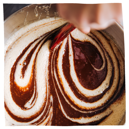 A close-up of a hand holding a red spatula and swirling dark chocolate and cream-colored brownie batter in a round metal bowl.