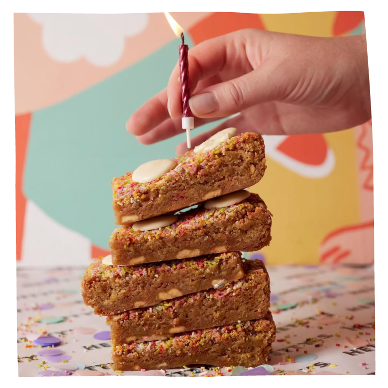 A stack of four brownies with a hand holding a lit candle above the stack against a colourful abstract background.