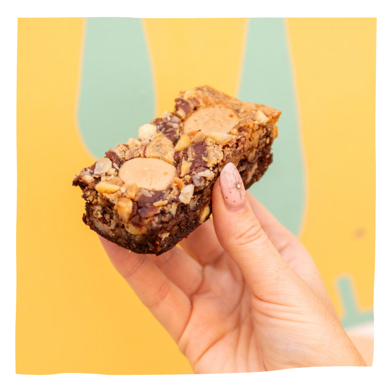 A hand holding a thick, chewy brownie, set against a bright yellow background with abstract green shapes.