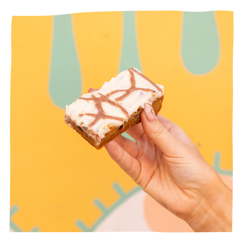 A hand holding a square slice of  brownie with white frosting and a brown crisscross drizzle, set against a bright yellow background with abstract mint green shapes.