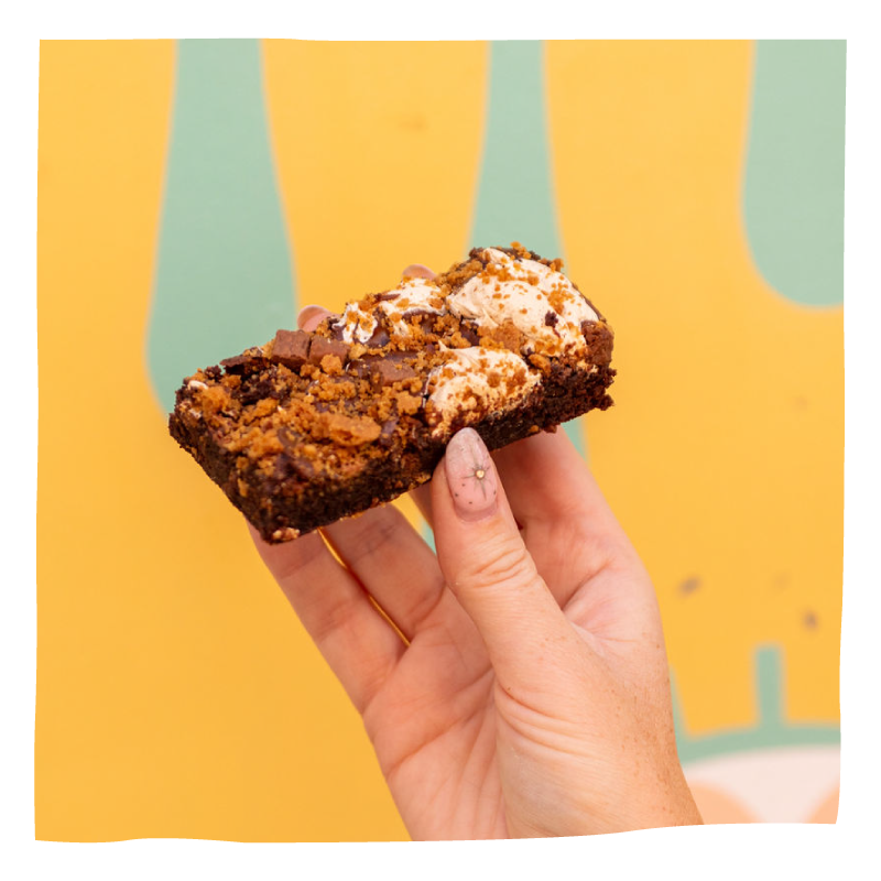 A hand holding a chocolate brownie, set against a bright yellow background with abstract green shapes.