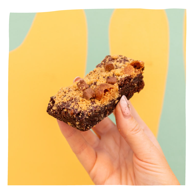 A hand holding a brownie, set against a bright yellow and mint green abstract background.
