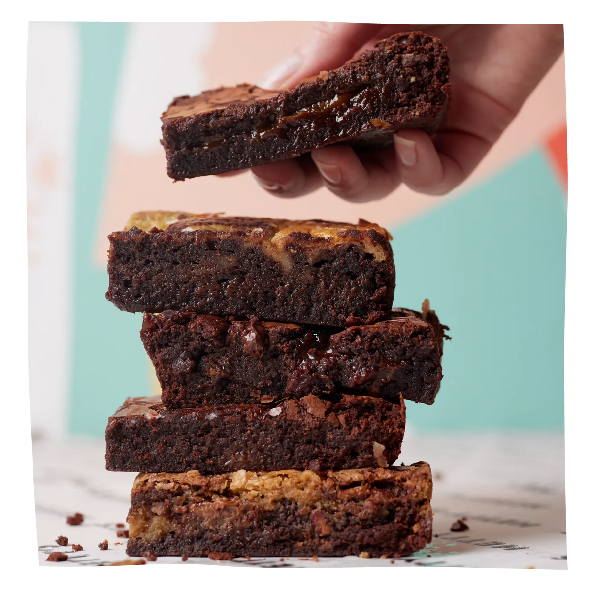 Four rich chocolate brownies stacked on a marble surface, with a hand lifting the top brownie slice to show its thick, gooey layer. The background is a soft, blurred pastel mix of mint green, pink, and beige.
