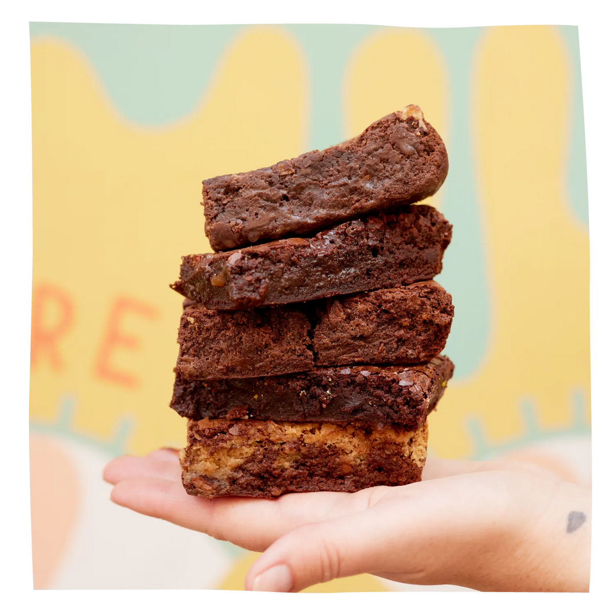 A hand holding a stack of five rich, dark brown chocolate brownies against a colorful, abstract pastel yellow and mint green background.