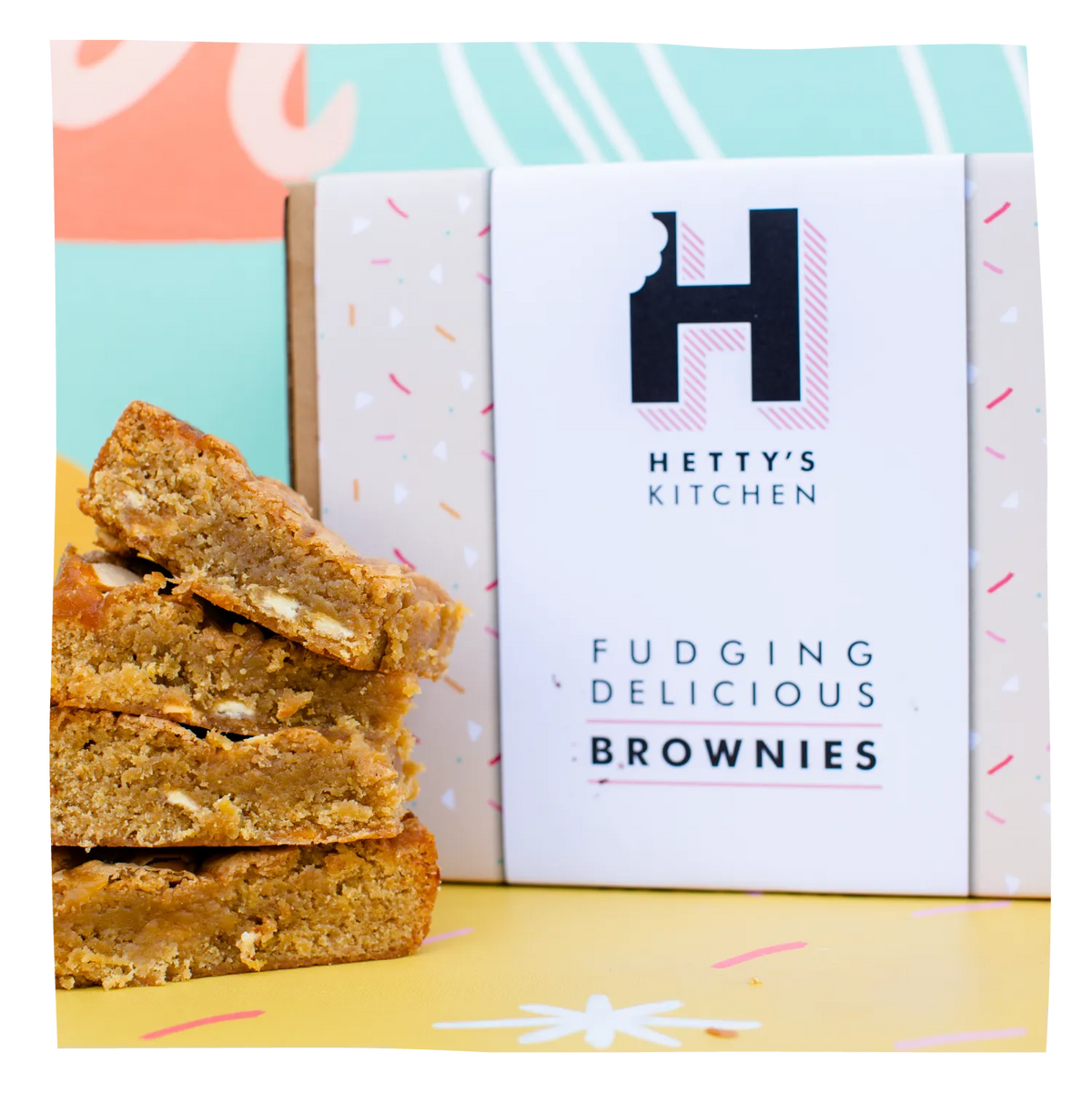 A box of Hetty's Kitchen Fudging Delicious Brownies sits next to a stack of three golden-brown brownie bars on a yellow surface with a colorful pastel background.