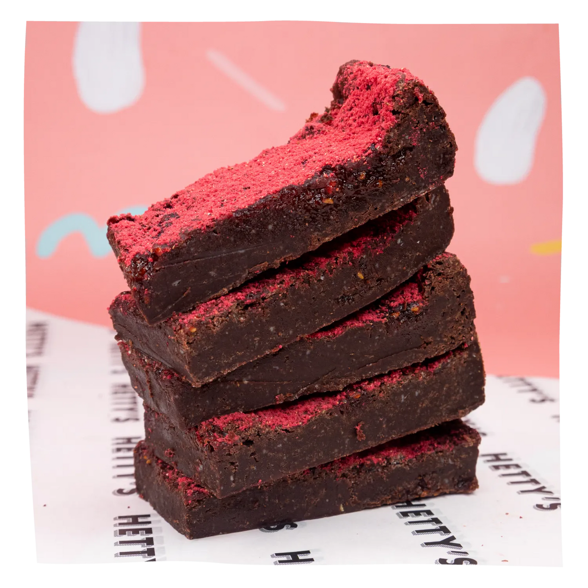 A stack of four square chocolate brownies topped with bright pink powder, set on a white paper surface with a pink background.