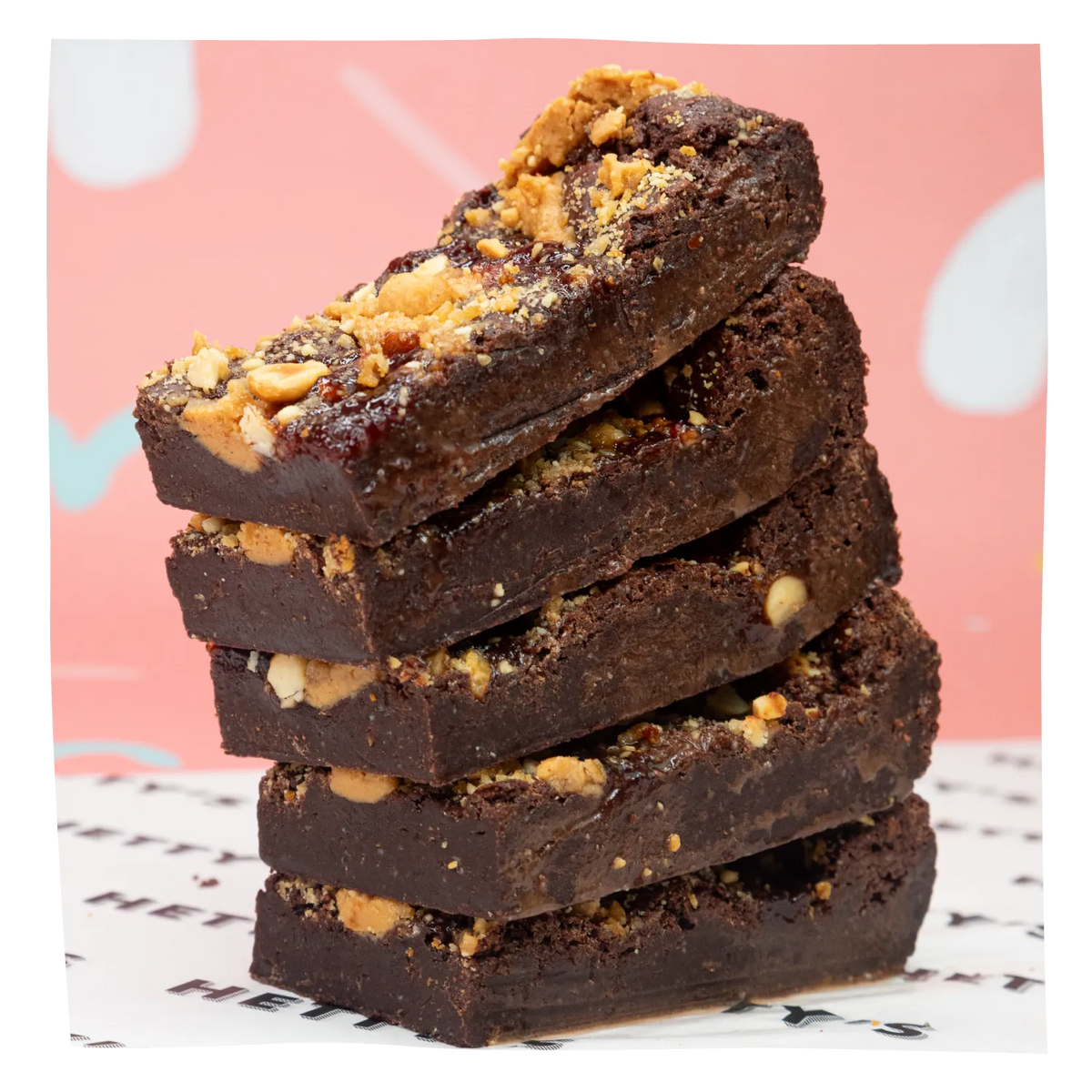 A stack of five thick chocolate brownies topped with chopped nuts, set against a pink background with white abstract shapes.