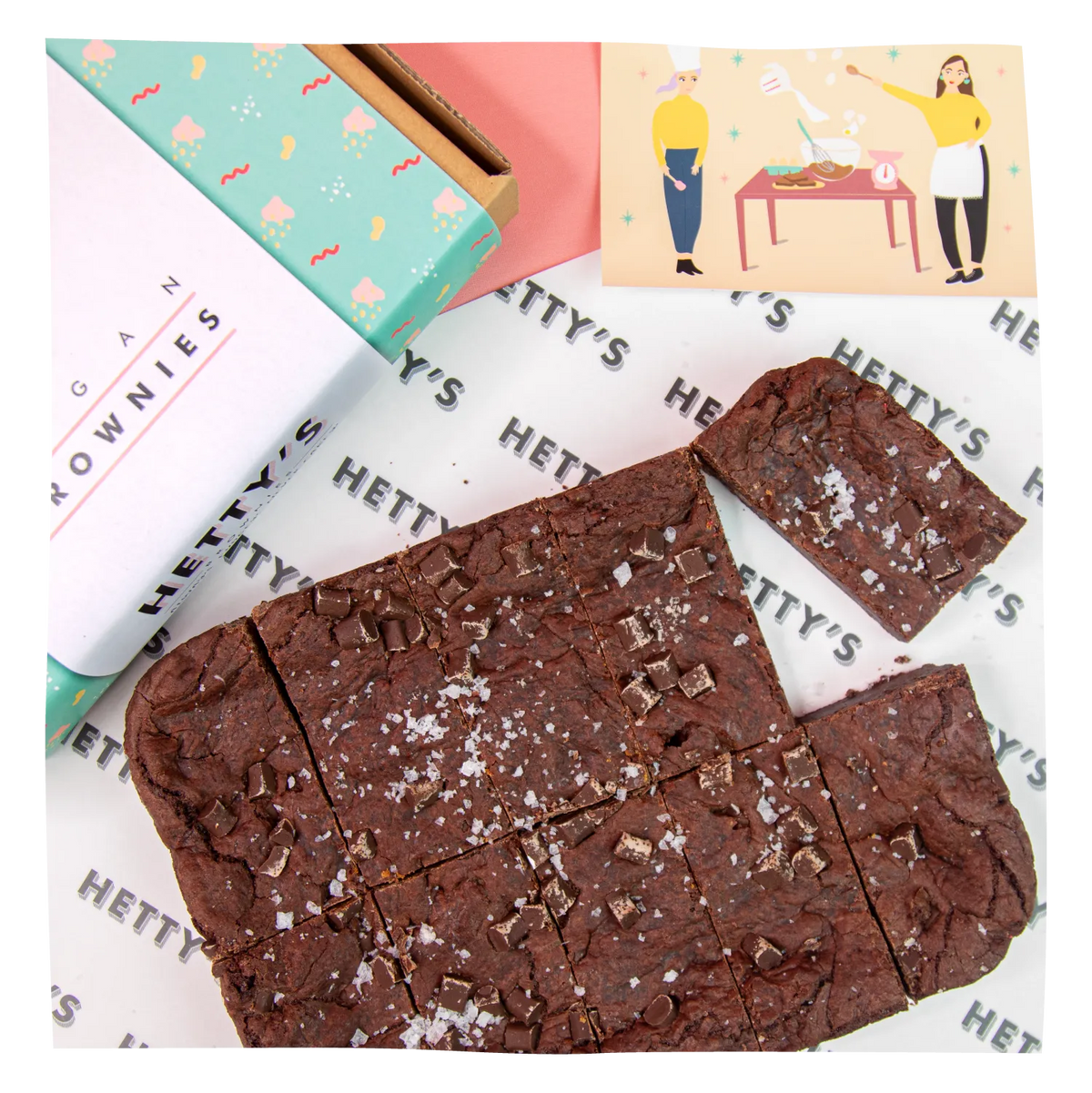Chocolate brownies topped with chocolate chips and coarse salt, arranged on branded paper with a box and illustrated card visible in the background.