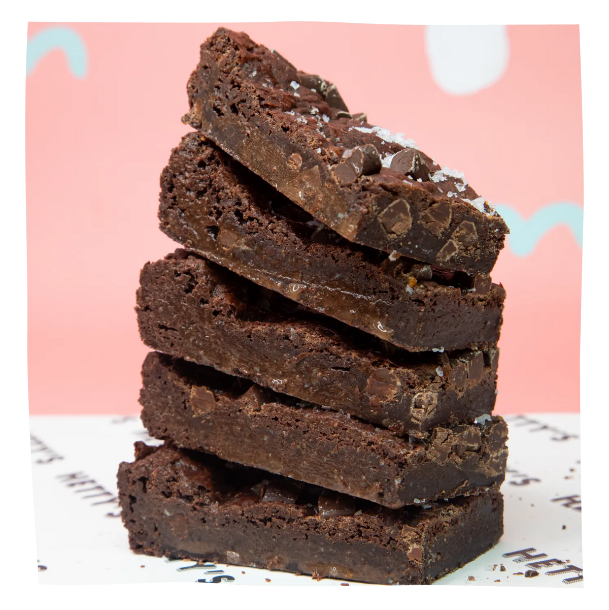 A stack of five thick chocolate brownies with chocolate chips and a sprinkle of coarse sea salt, set against a pink background with a white patterned surface.