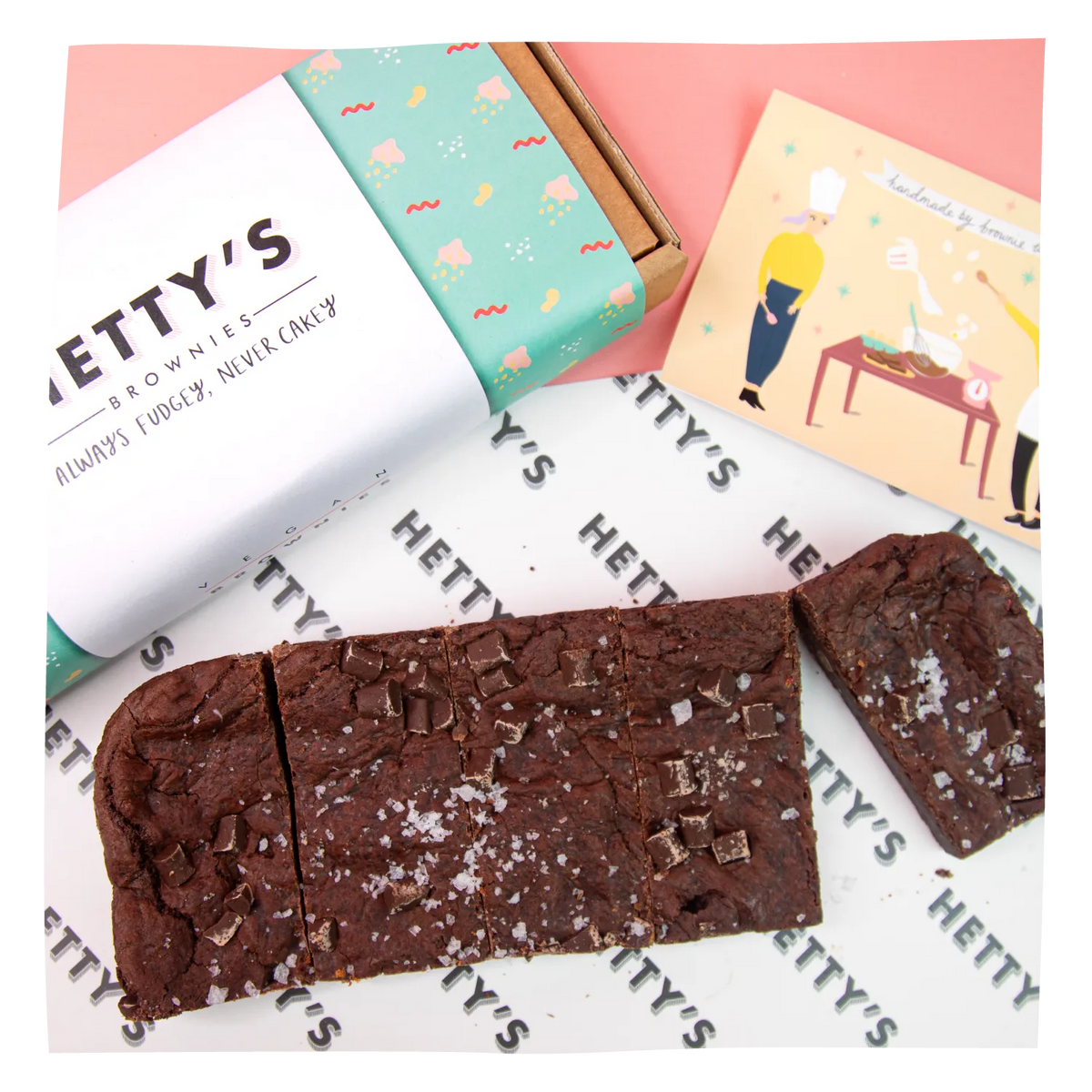 Alarge brownie bar broken in half to show the texture and toppings, alongside a branded box and a small illustrated card on a pink and white background.