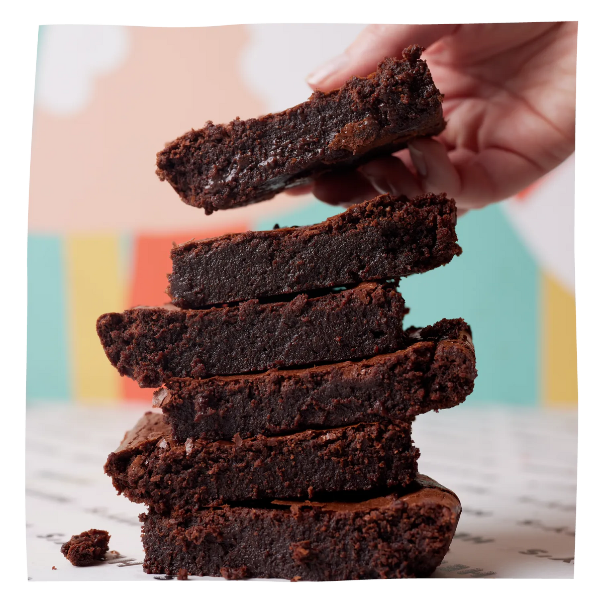 A stack of five thick chocolate brownies with the top brownie being lifted by a hand, set against a softly blurred pastel background.