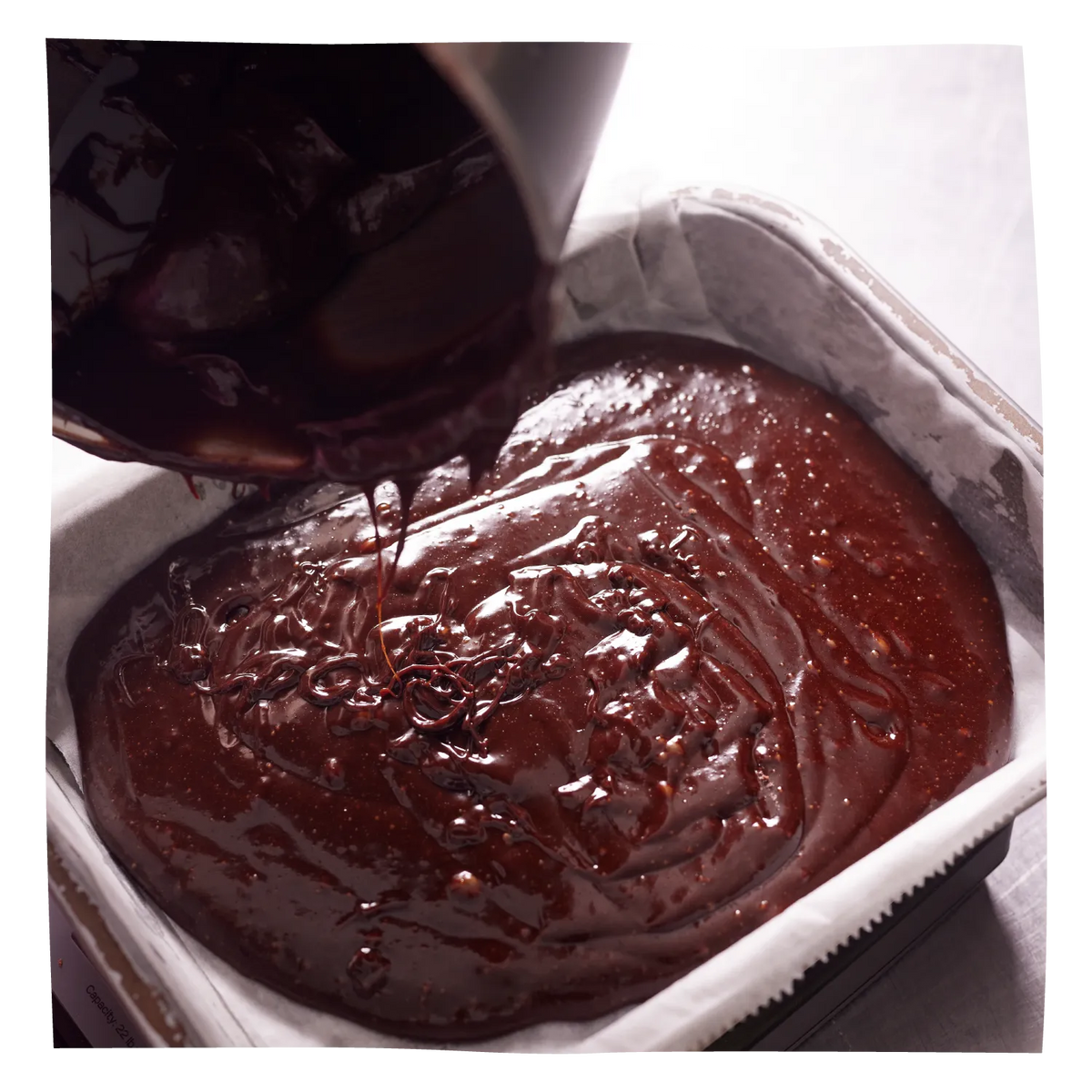 Rich, glossy chocolate brownie batter being poured from a metal pot into a square baking pan lined with white parchment paper.