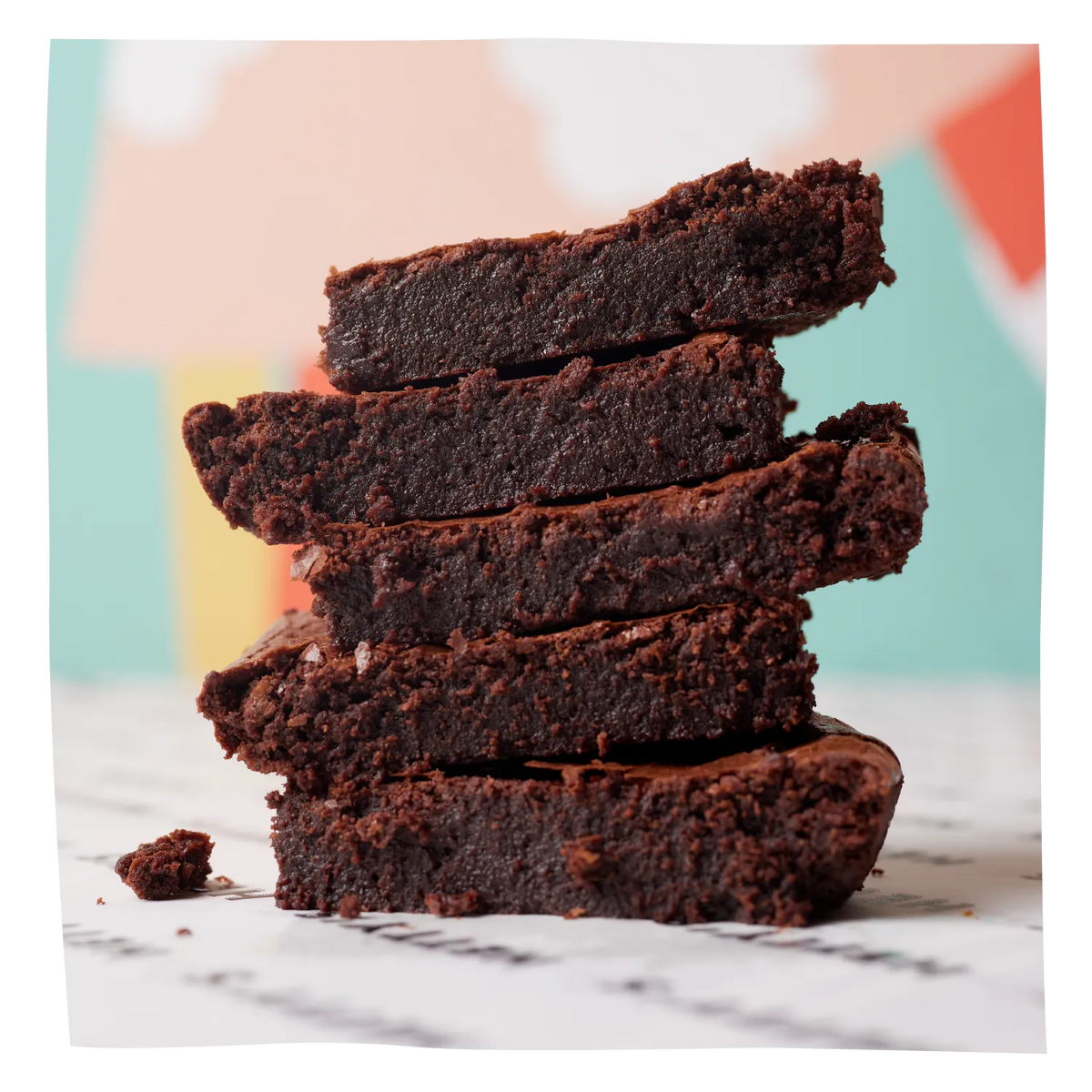 A stack of five thick, rich chocolate brownies with a crumbly, slightly glossy top, set against a softly blurred pastel background.