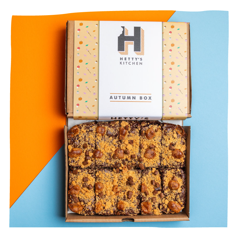 An open cardboard box of Hetty's Kitchen 'Autumn Box' treats, showing brownies, set against a split orange and light blue background.