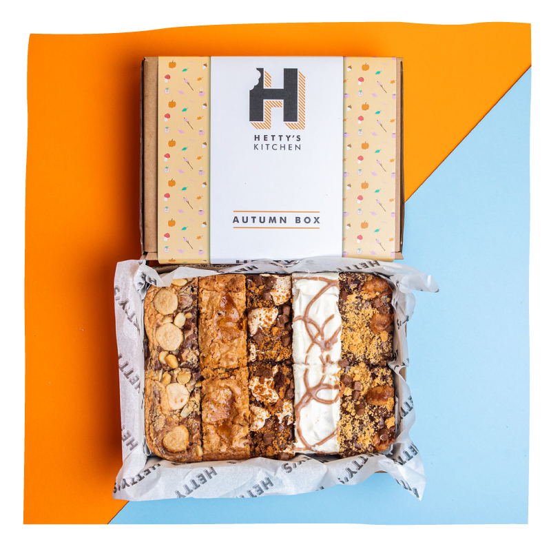 An overhead flat-lay of a boxed food product on a split orange and light blue background. The top shows the branded lid reading 'Hetty's Kitchen' and 'Autumn Box,' while the open bottom reveals assorted baked brownies wrapped in branded paper.