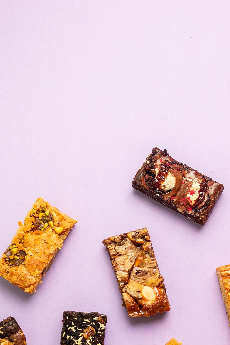 Several assorted brownies are scattered on a light lavender background, with some bars cropped at the edges of the frame.