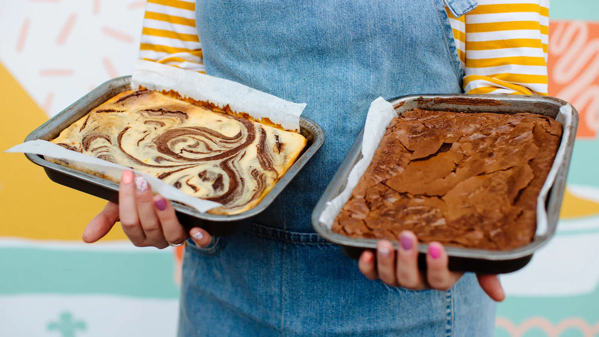 A person wearing denim overalls over a yellow-and-white striped shirt holds two baking pans of brownies. 