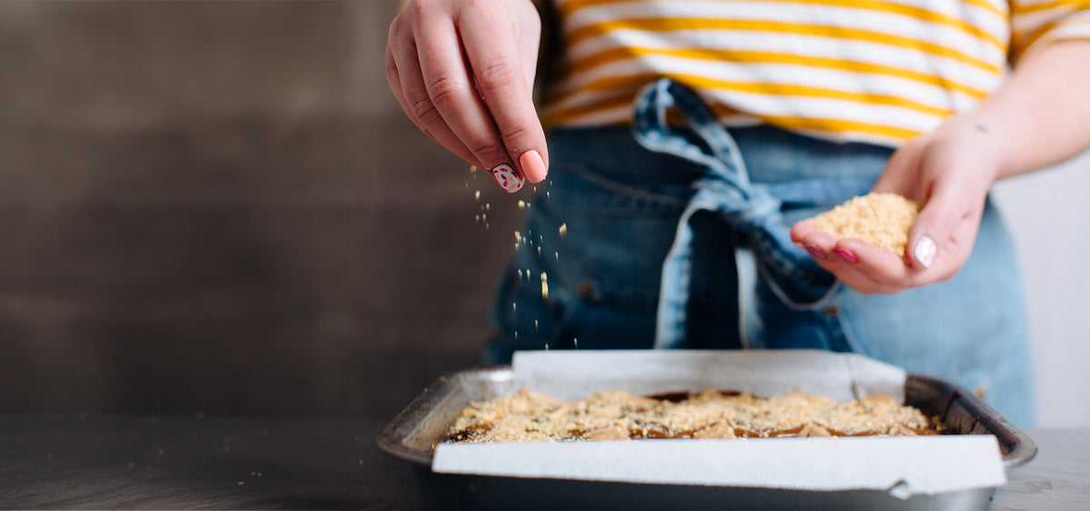 A person wearing a yellow and white striped shirt and blue apron sprinkles over brownies in a metal baking pan lined with parchment paper.