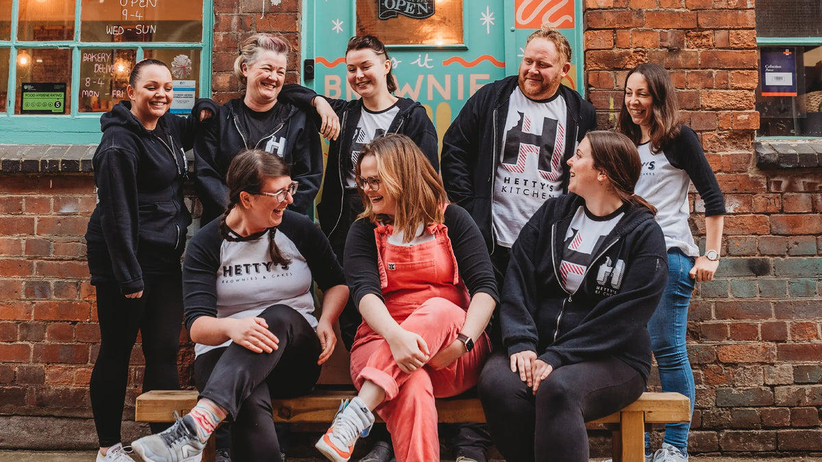 A group of eight people posing together on a wooden bench in front of a brick building with a teal door.
