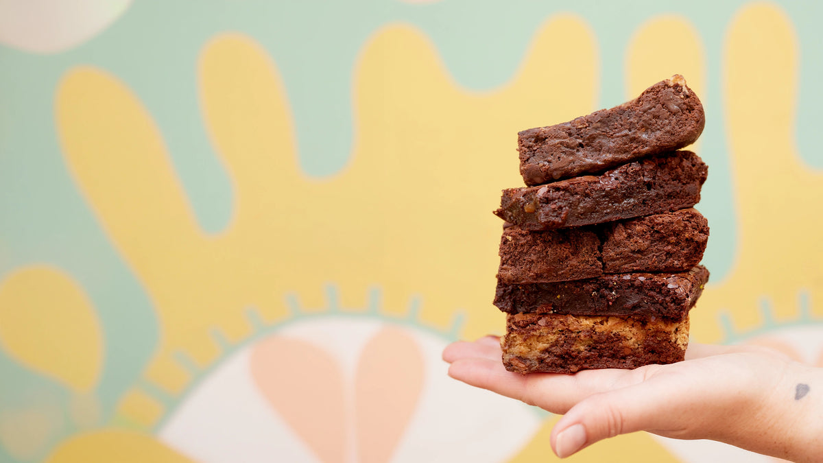 A hand holding a stack of five chocolate brownies in front of a colorful, abstract background with yellow and mint green shapes.